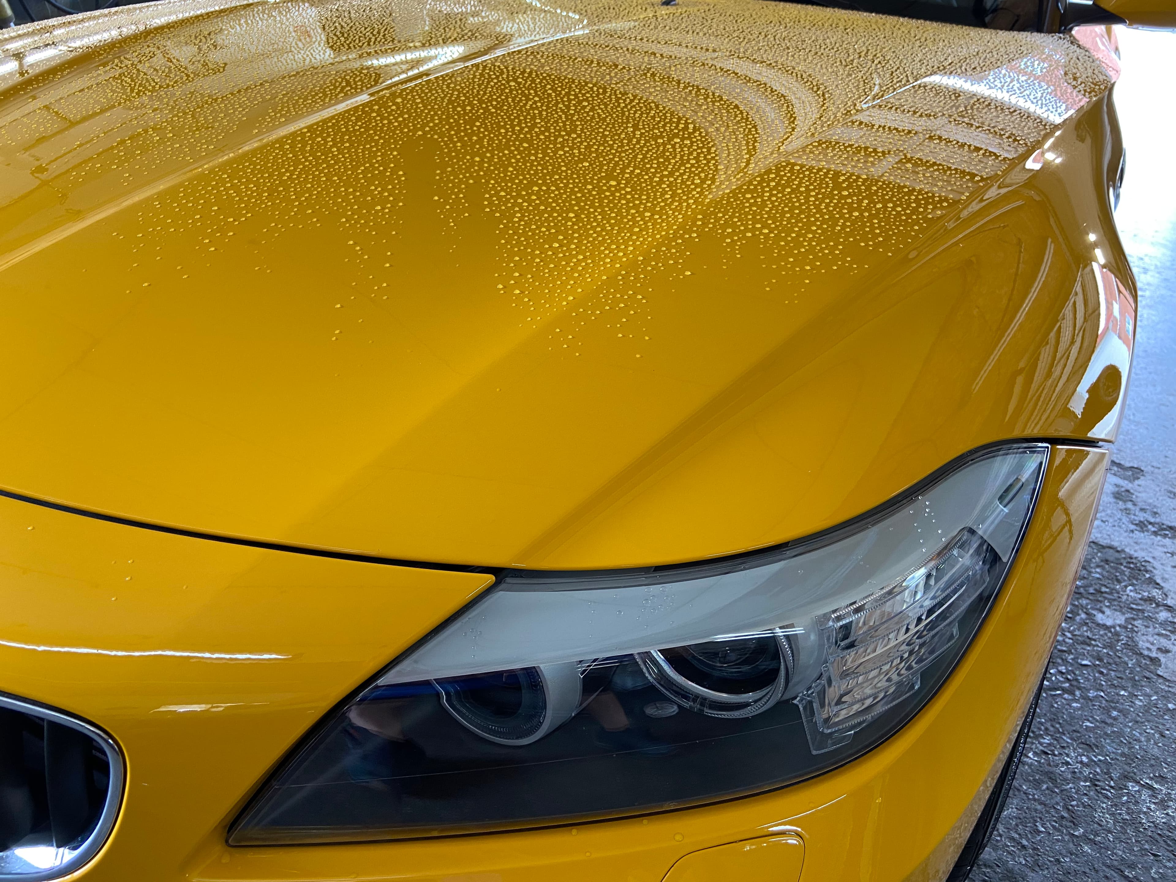 Close-up of water droplets beading on a bright yellow car hood and sleek headlight.